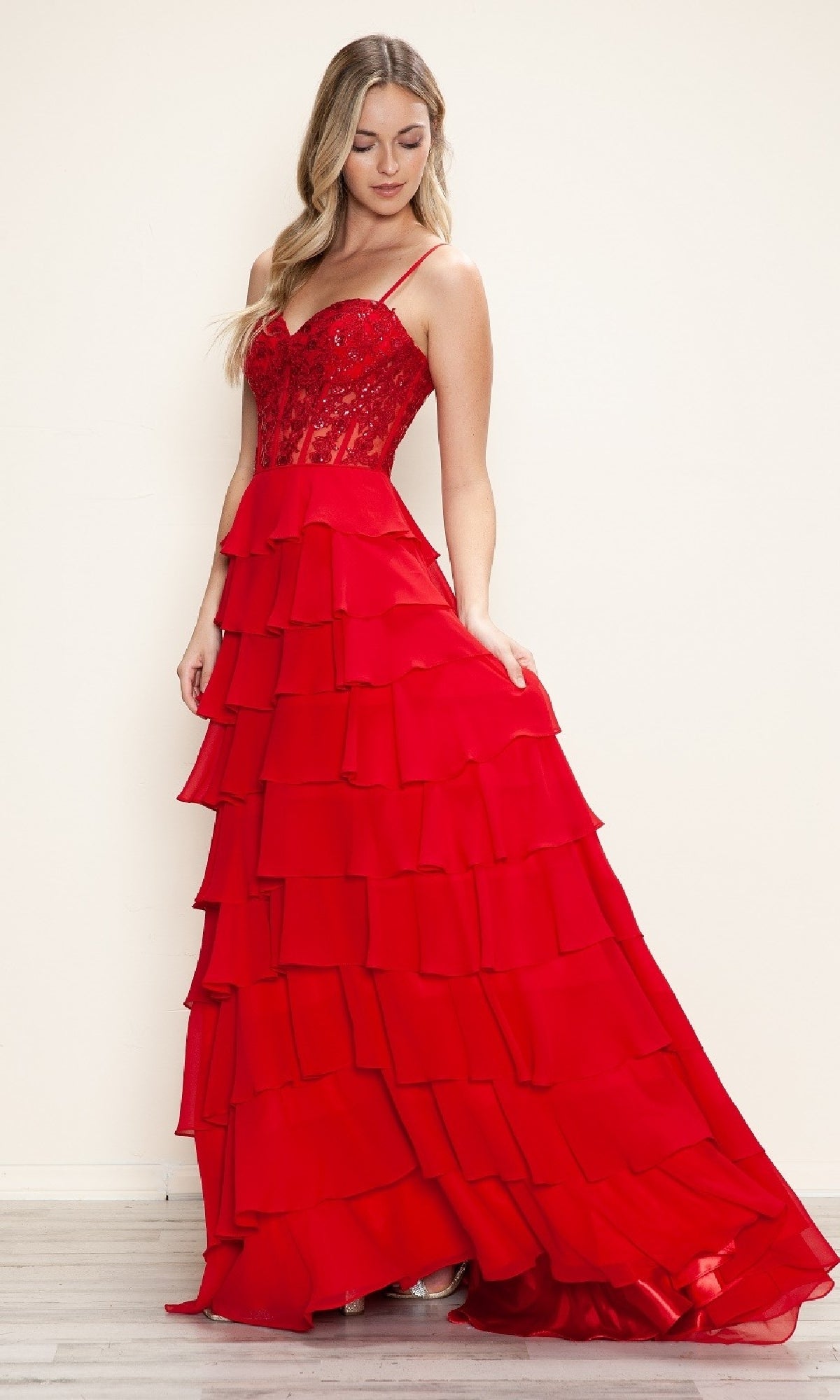 Woman wearing the Long Tiered Corset Prom Dress: Poly USA 9698, a red A-line gown with a fitted corset bodice and spaghetti straps, standing against a plain light background.
